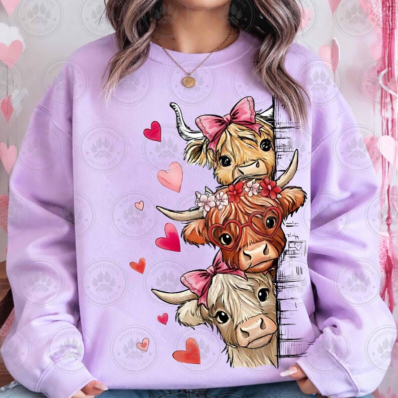 Puede incluir: Una sudadera lila claro con un gr&aacute;fico de tres vacas de las Tierras Altas adornadas con lazos rosas y gafas en forma de coraz&oacute;n. Corazones rojos y rosas est&aacute;n esparcidos alrededor de las vacas. Las vacas se asoman detr&aacute;s de una pared de ladrillos.