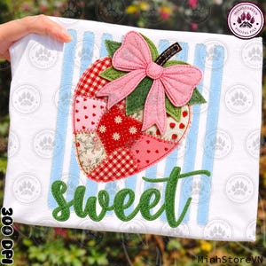 May include: White t-shirt featuring a patchwork strawberry design with a pink bow and green leaves. The strawberry is composed of various red and pink patterned fabrics. The word "sweet" is written in green script below the strawberry.