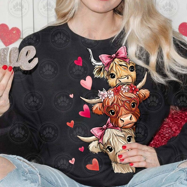 Puede incluir: Sudadera negra con un dise&ntilde;o apilado de tres vacas de las Tierras Altas de dibujos animados. Las vacas tienen lazos rosas, gafas en forma de coraz&oacute;n y detalles florales. Corazones rosas y rojos est&aacute;n dispersos, creando un tema de San Valent&iacute;n.