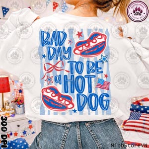 Può includere: Felpa bianca con un design patriottico. La parte posteriore presenta la scritta "BAD DAY TO BE A HOT DOG" in blu, con illustrazioni di hot dog rossi e blu, stelle e una bandiera americana. Un design festoso e divertente.