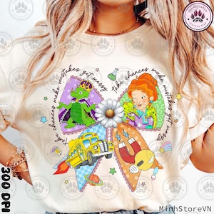 Può includere: T-shirt color crema con un design grafico colorato. Il design include una forma a fiocco con illustrazioni di un autobus scolastico, un drago, un'insegnante e un magnete. Il testo sulla maglietta recita "Take chances, make mistakes, get messy."