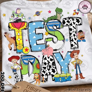 Puede incluir: Camiseta blanca con las palabras "TEST DAY" en una fuente lúdica, rodeada de personajes de dibujos animados. El diseño incluye un cohete, estrellas y varios personajes de Toy Story. La camiseta tiene una etiqueta que dice "THRIFT STORE".