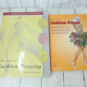 May include: Two softcover books titled The Art of Fashion Draping Second Edition and Fashion Prints. The first book has a yellow cover with a line drawing of a draped garment. The second book has an orange cover with an illustration of a person.