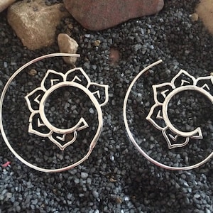 Puede incluir: Un par de pendientes en espiral de color plateado. Cada pendiente presenta un diseño floral en el centro de la espiral. Los pendientes se exhiben sobre un lecho de pequeñas piedras de color gris oscuro.