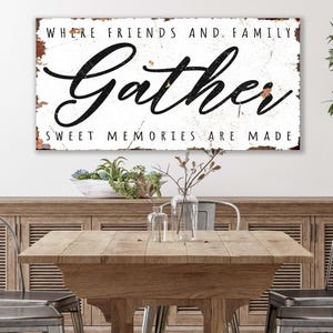 May include: A rectangular wooden sign with the words "Where Friends and Family Gather, Sweet Memories Are Made" in black script. The sign is mounted above a wooden table with metal chairs. A vase with greenery is on the right.