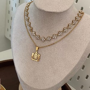 Puede incluir: Juego de collares dorados expuestos en un maniquí. El collar superior presenta un diseño floral con piedras preciosas transparentes. El collar inferior tiene un colgante con un diseño ornamentado.