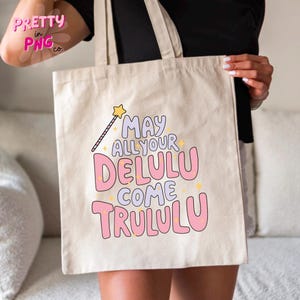 Pode incluir: Uma bolsa tote de lona branca com texto rosa e roxo que diz "May all your Delulu come Trululu". Uma estrela dourada e uma varinha m&aacute;gica est&atilde;o acima do texto.