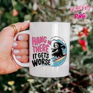 May include: White ceramic mug with the text "HANG IN THERE IT GETS WORSE" in pink and black. The mug features a cartoon grim reaper surfing a wave. The mug has a white handle and is held by a hand.