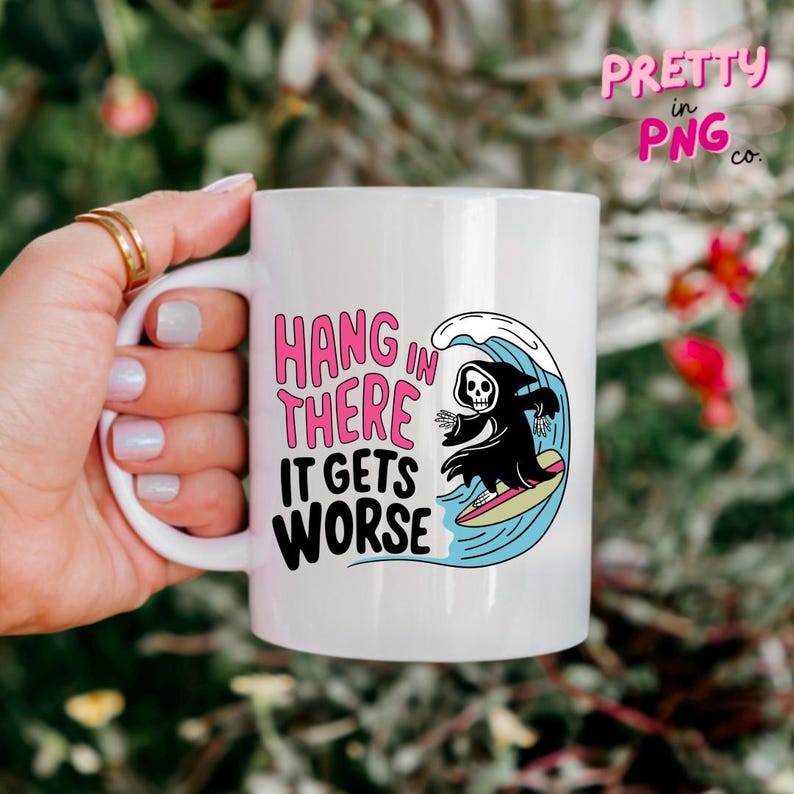 May include: White ceramic mug with the text "HANG IN THERE IT GETS WORSE" in pink and black. The mug features a cartoon grim reaper surfing a wave. The mug has a white handle and is held by a hand.