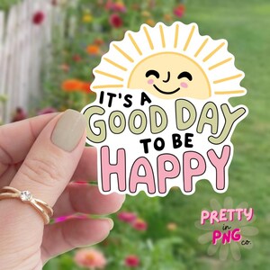 May include: A sticker with a smiling sun and the text "It's a good day to be happy" in black, green, and pink. The sticker has a white border and the words "Pretty in PNG co." in pink.