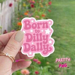 May include: A pink sticker with the words "Born to Dilly Dally" in a playful font, accented with white stars. The sticker is held up against a blurred background of green grass and flowers.