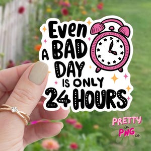 May include: White sticker with black text that reads "Even a bad day is only 24 hours". A pink alarm clock with black hands and black stars is next to the text. The sticker has a white border and a glittery background.