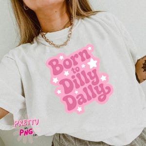 May include: White t-shirt with a pink graphic that reads "Born to Dilly Dally" with white star accents. The shirt also has the text "Pretty in PNG co." in the bottom left corner. The shirt is a casual style.