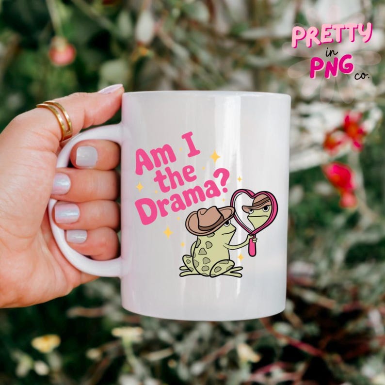Pode incluir: Caneca de cer&acirc;mica branca com um design de sapo rosa e verde. O sapo est&aacute; usando um chap&eacute;u de cowboy e segurando um espelho em forma de cora&ccedil;&atilde;o. O texto "Am I the Drama?" est&aacute; impresso em rosa na caneca.