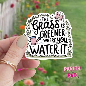 May include: A white sticker with a green grass border and a pink butterfly. The sticker features the text "*THE GRASS IS GREENER WHERE you WATER IT*" with a pink watering can and a white star.