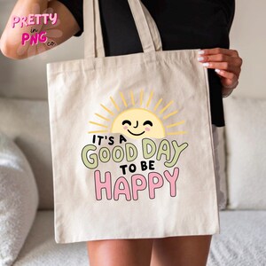 May include: Beige tote bag with the text "It's a good day to be happy" in green, pink, and black. A smiling sun graphic is above the text. The bag has two handles and is held by a person.