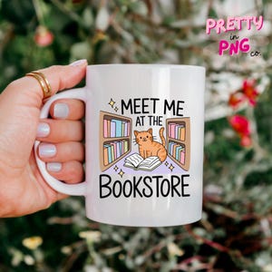 May include: White ceramic mug featuring the text "Meet Me at the Bookstore" with a cartoon cat and bookshelves design. The mug has a curved handle and is held by a hand with gold rings.