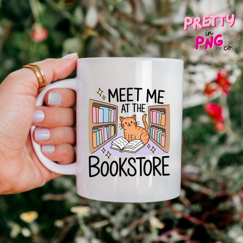 May include: White ceramic mug featuring the text "Meet Me at the Bookstore" with a cartoon cat and bookshelves design. The mug has a curved handle and is held by a hand with gold rings.