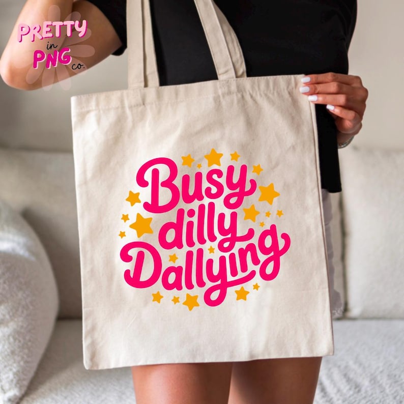 以下が含まれることがあります： ピンク色の筆記体と黄色の星で「Busy Dilly Dallying」と書かれたベージュのトートバッグ。バッグには2つのハンドルがあり、人が持っています。「Pretty in PNG co.」というテキストが左上にあります。