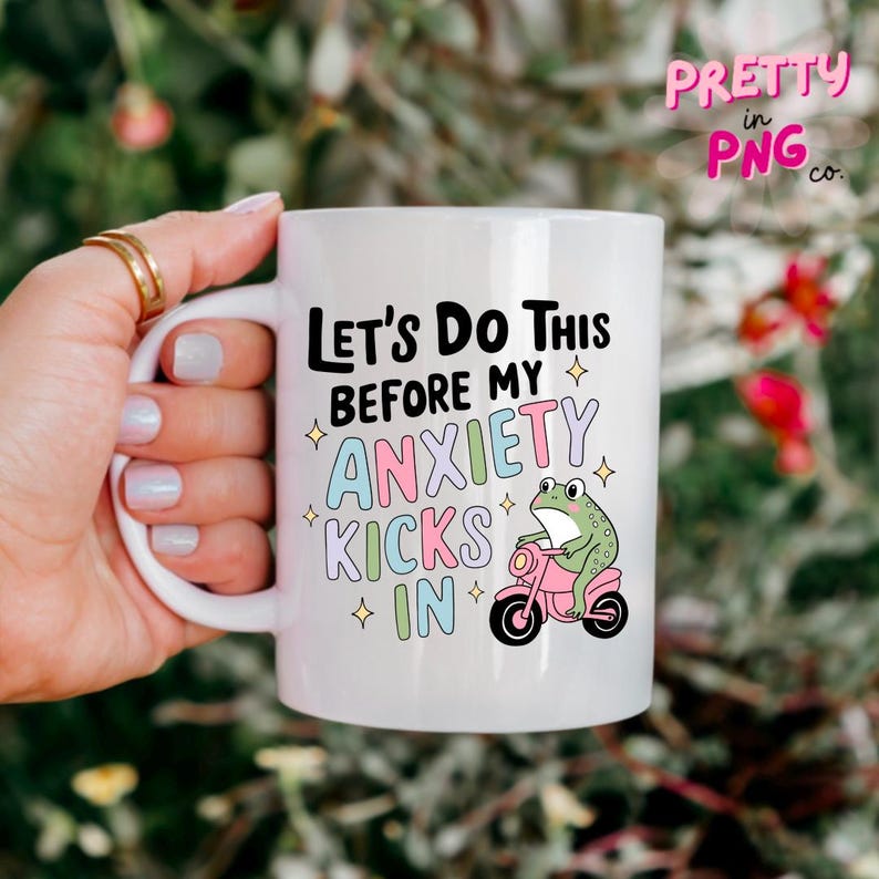 K&ouml;nnte beinhalten: Eine wei&szlig;e Keramiktasse mit einem schwarzen und rosafarbenen Design. Die Tasse zeigt eine Comic-Frosch, der ein Motorrad f&auml;hrt, mit dem Text "Let's do this before my anxiety kicks in".