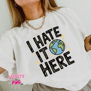 May include: White t-shirt featuring the text "I HATE IT HERE" in black, green, and blue, with a globe graphic and yellow star accents. The shirt also has the text "PRETTY in PNG co."
