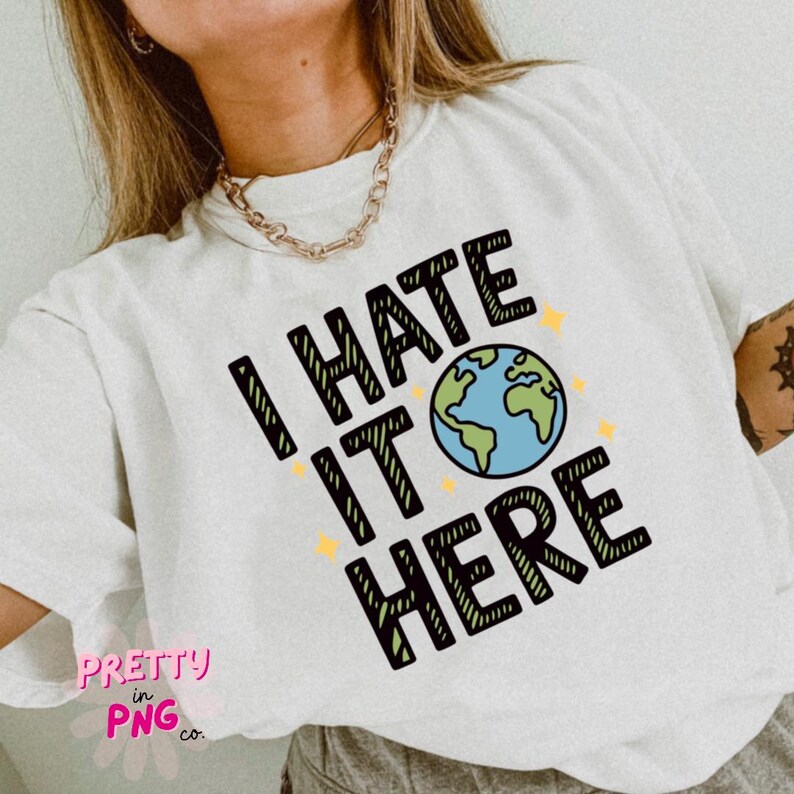 May include: White t-shirt featuring the text "I HATE IT HERE" in black, green, and blue, with a globe graphic and yellow star accents. The shirt also has the text "PRETTY in PNG co."