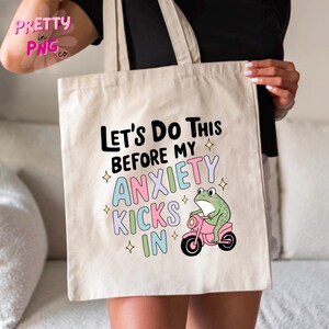 K&ouml;nnte beinhalten: Eine wei&szlig;e Canvas-Tragetasche mit dem Text "Let's do this before my anxiety kicks in" in Schwarz. Darunter befindet sich ein Cartoon-Frosch, der ein rosa Motorrad f&auml;hrt.