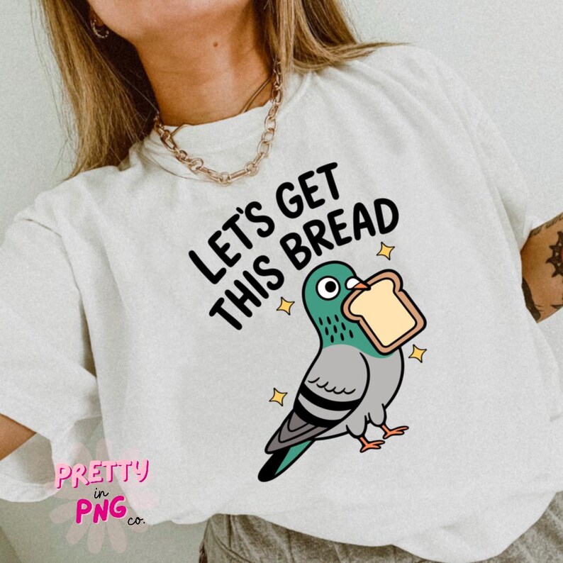Może przedstawiać: Biała koszulka z napisem "LET'S GET THIS BREAD" i rysunkiem gołębia trzymającego kromkę chleba. Gołąb jest szary, turkusowy i czarny z ż&oacute;łtymi gwiazdkami. Koszulka ma okrągły dekolt.