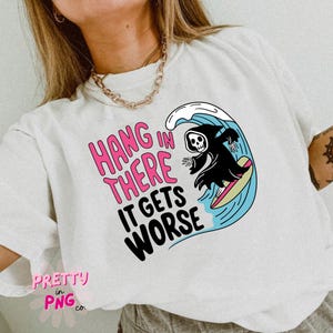 May include: White t-shirt featuring the text "Hang in There It Gets Worse" in pink and black. A cartoon grim reaper surfs a wave. The shirt has a relaxed fit.