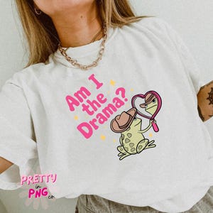 Pode incluir: Camiseta branca com texto rosa que diz "Am I the Drama?" e um sapo de desenho animado usando um chap&eacute;u de cowboy e olhando para um espelho em forma de cora&ccedil;&atilde;o. O sapo &eacute; verde com manchas marrons.
