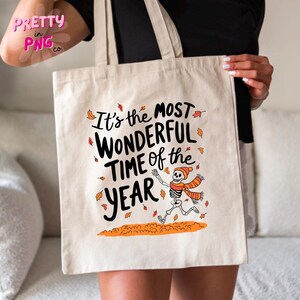 May include: A white tote bag with a black and orange design. The bag features a skeleton wearing a red and orange scarf and the text "It's the most wonderful time of the year".