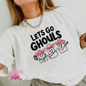 Pode incluir: Camiseta branca com o texto "Lets Go Ghouls" em preto. Tr&ecirc;s fantasmas de desenho animado usando chap&eacute;us de cowboy rosa est&atilde;o abaixo do texto.