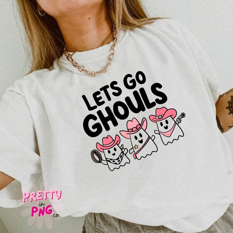 Pode incluir: Camiseta branca com o texto "Lets Go Ghouls" em preto. Tr&ecirc;s fantasmas de desenho animado usando chap&eacute;us de cowboy rosa est&atilde;o abaixo do texto.