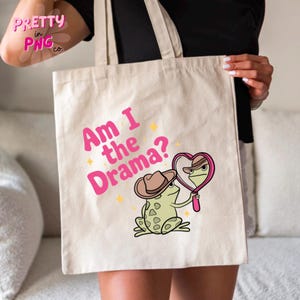 Pode incluir: Uma bolsa tote de lona branca com texto rosa que diz "Am I the Drama?" e um sapo de desenho animado usando um chap&eacute;u de cowboy olhando para si mesmo em um espelho em forma de cora&ccedil;&atilde;o.