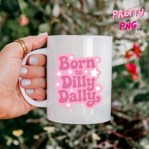May include: White ceramic mug with a pink graphic that reads "Born to Dilly Dally" with star accents. The mug also has the text "Pretty in PNG co." in pink. The mug is held by a hand with gold rings.