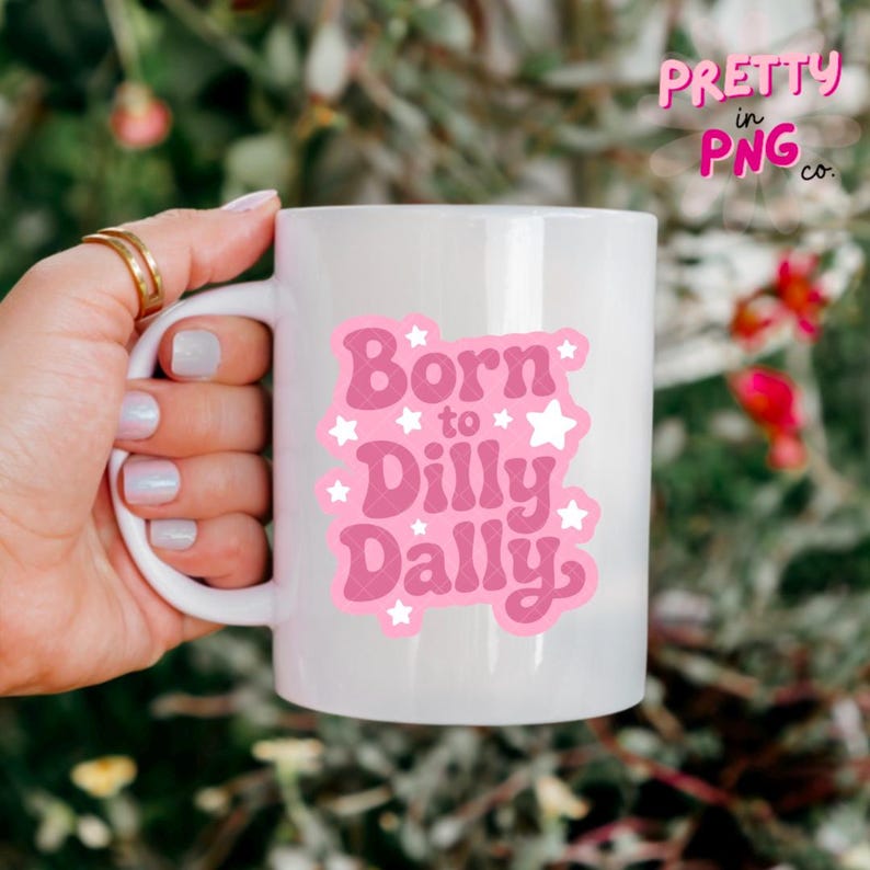 May include: White ceramic mug with a pink graphic that reads "Born to Dilly Dally" with star accents. The mug also has the text "Pretty in PNG co." in pink. The mug is held by a hand with gold rings.