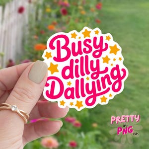 以下が含まれることがあります： ピンク色の筆記体で「Busy dilly Dallying」と書かれた白いステッカー。オレンジ色の星が散りばめられています。ステッカーは、金の指輪をつけた手によって持たれており、背景にはぼやけた花の模様があります。