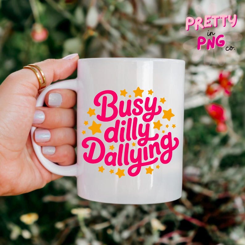 以下が含まれることがあります： ピンク色の文字と黄色の星で「Busy dilly Dallying」と書かれた白いセラミックマグ。 金色の指輪と薄いピンクのマニキュアを塗った手でマグカップを持っています。 背景は緑と赤の葉でぼやけています。