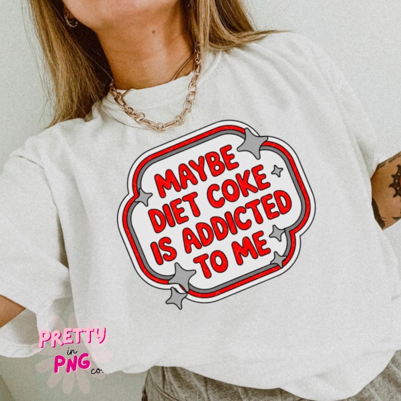 May include: White t-shirt with a graphic that reads "MAYBE DIET COKE IS ADDICTED TO ME" in red and white, outlined in gray and red. The design is in a rounded rectangle shape with star accents. The shirt has a relaxed fit.