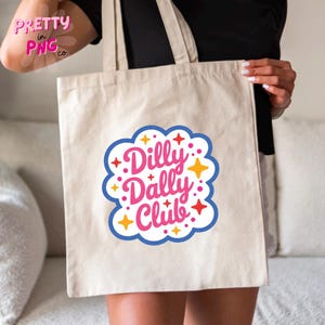 May include: A beige canvas tote bag featuring a central graphic with the text Dilly Dally Club in pink cursive lettering. The design is enclosed in a blue scalloped border and decorated with small yellow and red four-pointed star accents.