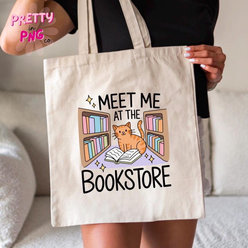 May include: Beige tote bag featuring the text "MEET ME AT THE BOOKSTORE" with an illustration of a cat reading in front of bookshelves. The cat is orange with black stripes. The bookshelves are filled with colorful books.