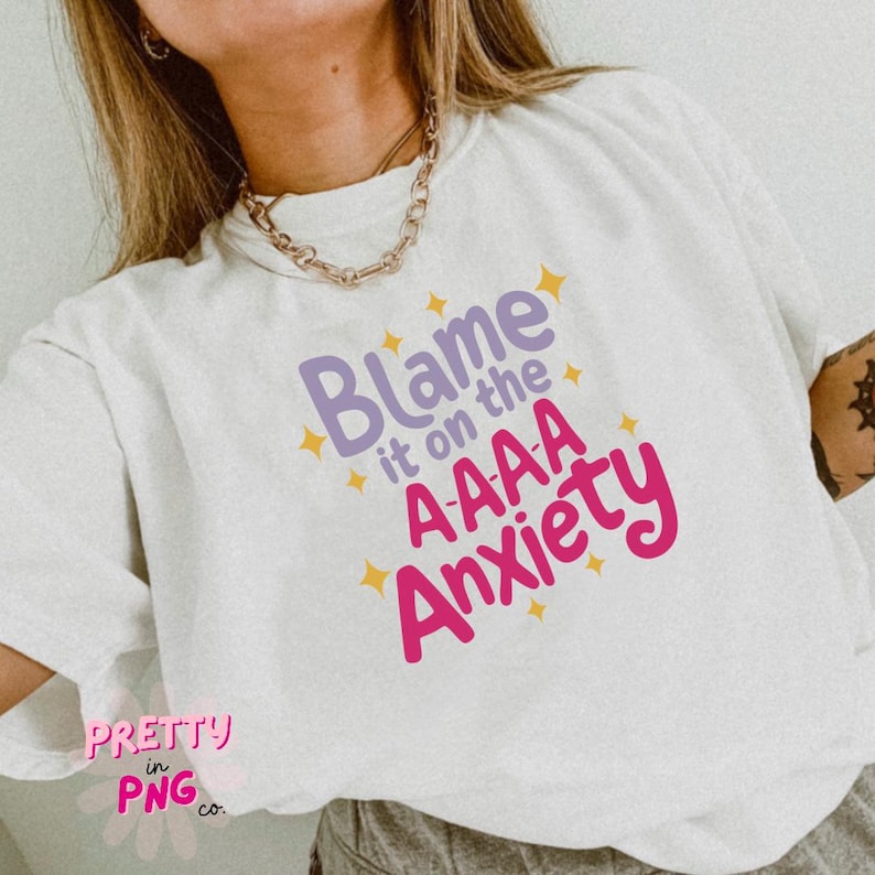 以下が含まれることがあります： 黄色い星が散りばめられた白いTシャツに、紫とピンクの文字で「Blame it on the A-AAA Anxiety」と書かれています。