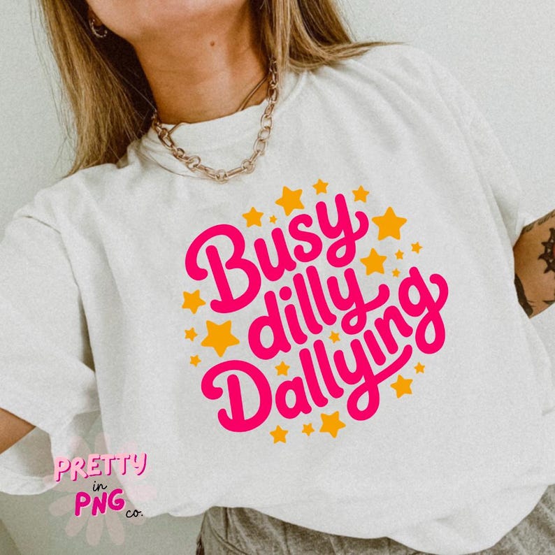 以下が含まれることがあります： 明るいピンク色のスクリプトで「Busy Dilly Dallying」というフレーズが書かれた白いTシャツ。オレンジ色の星が散りばめられています。また、ピンク色のスクリプトで「Pretty in PNG co.」という文字も書かれています。