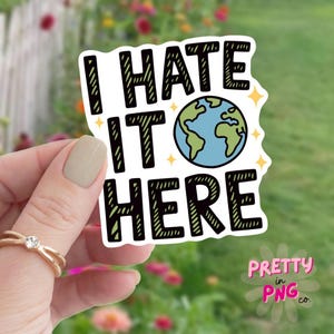 May include: A white sticker with the text "I HATE IT HERE" in black, featuring a blue and green globe graphic. The sticker has a decorative border with yellow starbursts and the words "Pretty in PNG co." in pink.