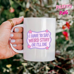May include: White ceramic mug with the text "I HAVE TO SAY WEIRD STUFF OR I'LL DIE" in a purple and pink design. The mug has a curved handle and is held by a person. The mug is from Pretty in PNG co.