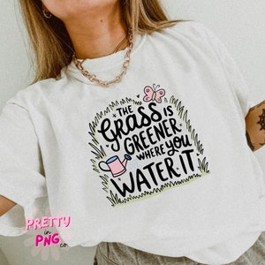 May include: White t-shirt with a green grass design and a pink watering can. The text reads "The grass is greener where you water it".