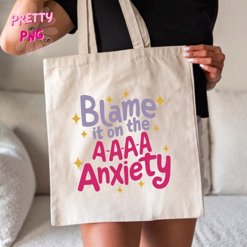 以下が含まれることがあります： ゴールドの星が散りばめられた、ピンクとパープルの文字で「Blame it on the A-A-A Anxiety」と書かれた白いキャンバス地のトートバッグです。