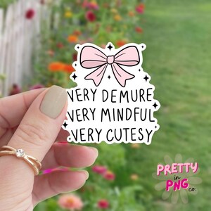 May include: A pink bow with a white outline and black stars. The text below the bow reads "Very Demure Very Mindful Very Cutesy".