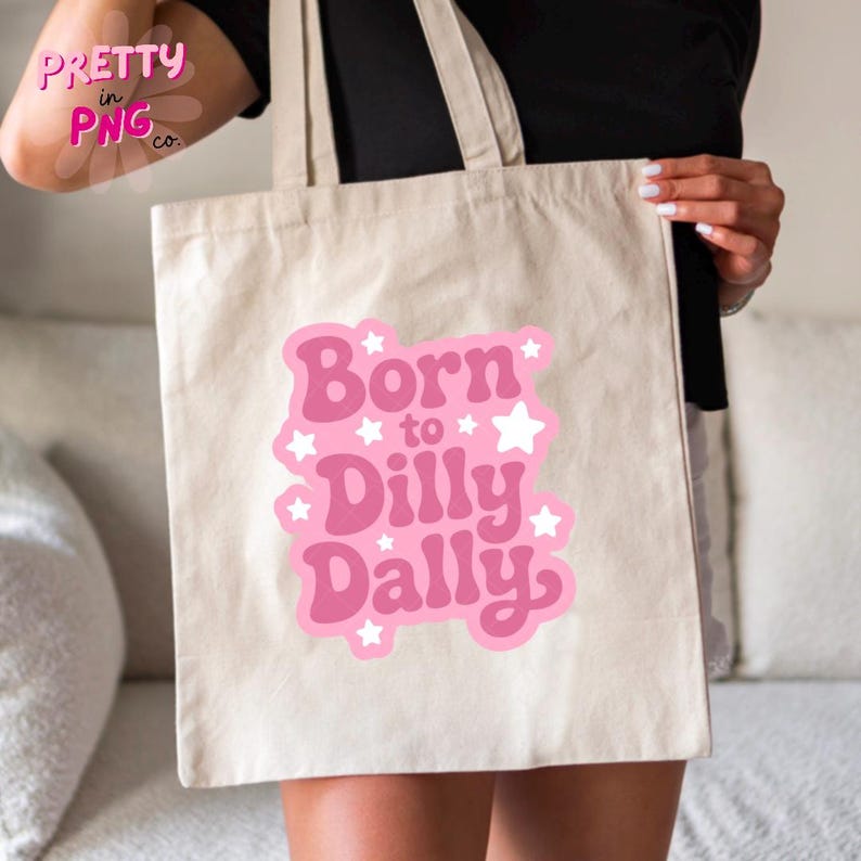 May include: A beige tote bag with the phrase "Born to Dilly Dally" in pink, with white star accents. The bag is held by a person.