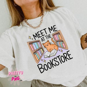 May include: White t-shirt with a graphic that says "MEET ME AT THE BOOKSTORE". The graphic features an orange cat reading a book in front of bookshelves with colorful books. The shirt has a relaxed fit.